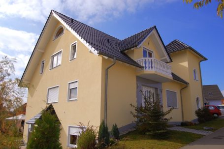 Der Hausbau Berater - Fachberater für Fertighäuser - Thomas Bärtle Helles Einfamilienhaus mit Balkon und grünem Vorgarten, umgeben von Bäumen.