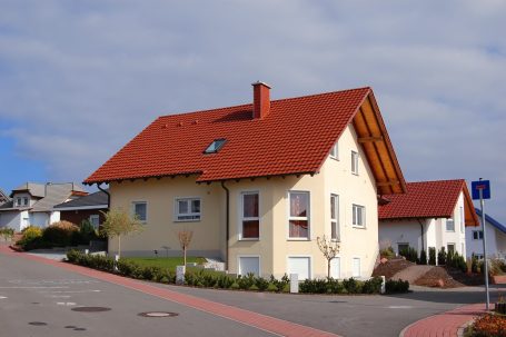Der Hausbau Berater - Fachberater für Fertighäuser - Thomas Bärtle Einfamilienhaus mit rotem Dach und umliegendem Garten in einer ruhigen Wohngegend.